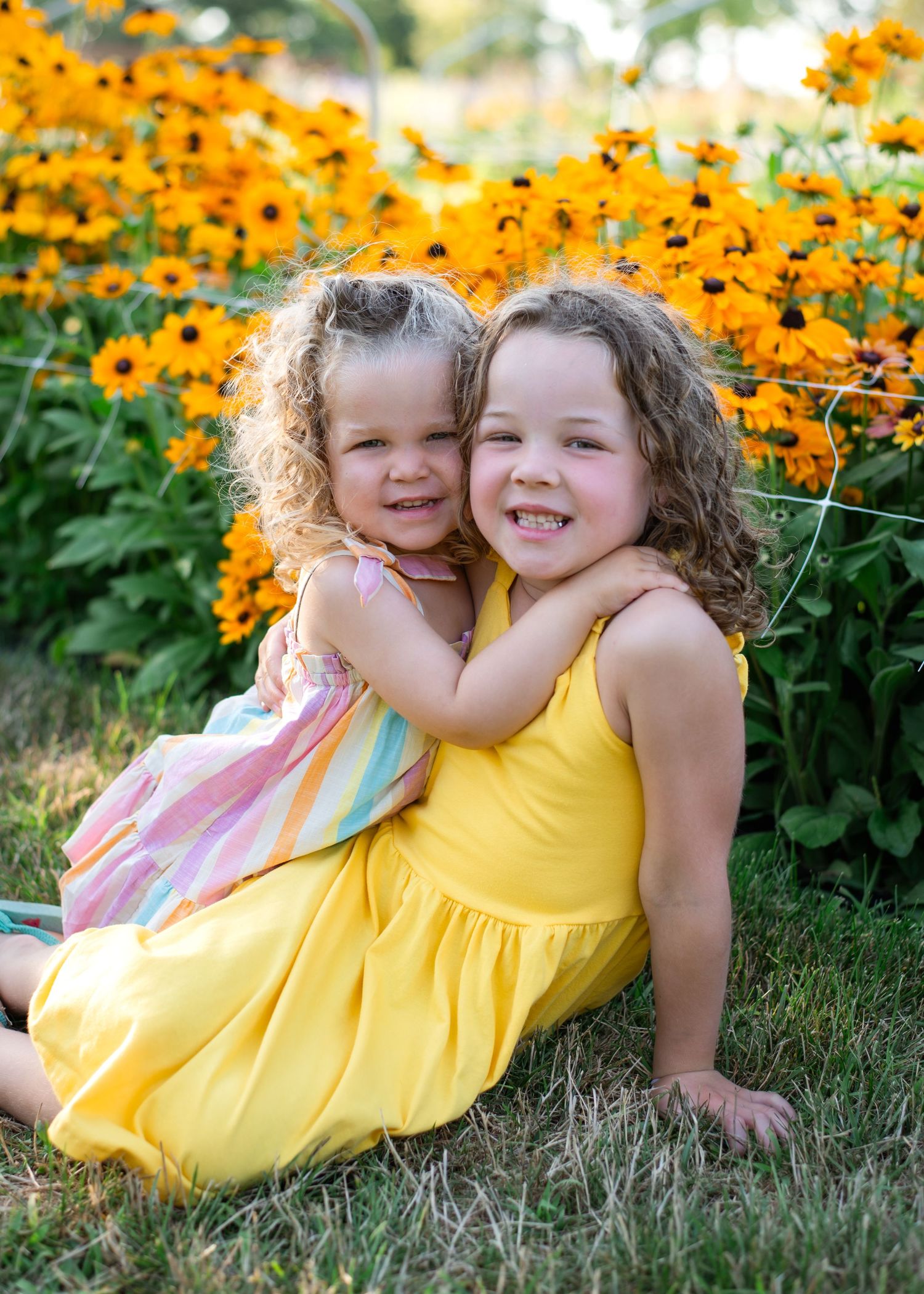Two children hugging and smiling in front of a vibrant garden of yellow Black-eyed Susan flowers on a summer day.