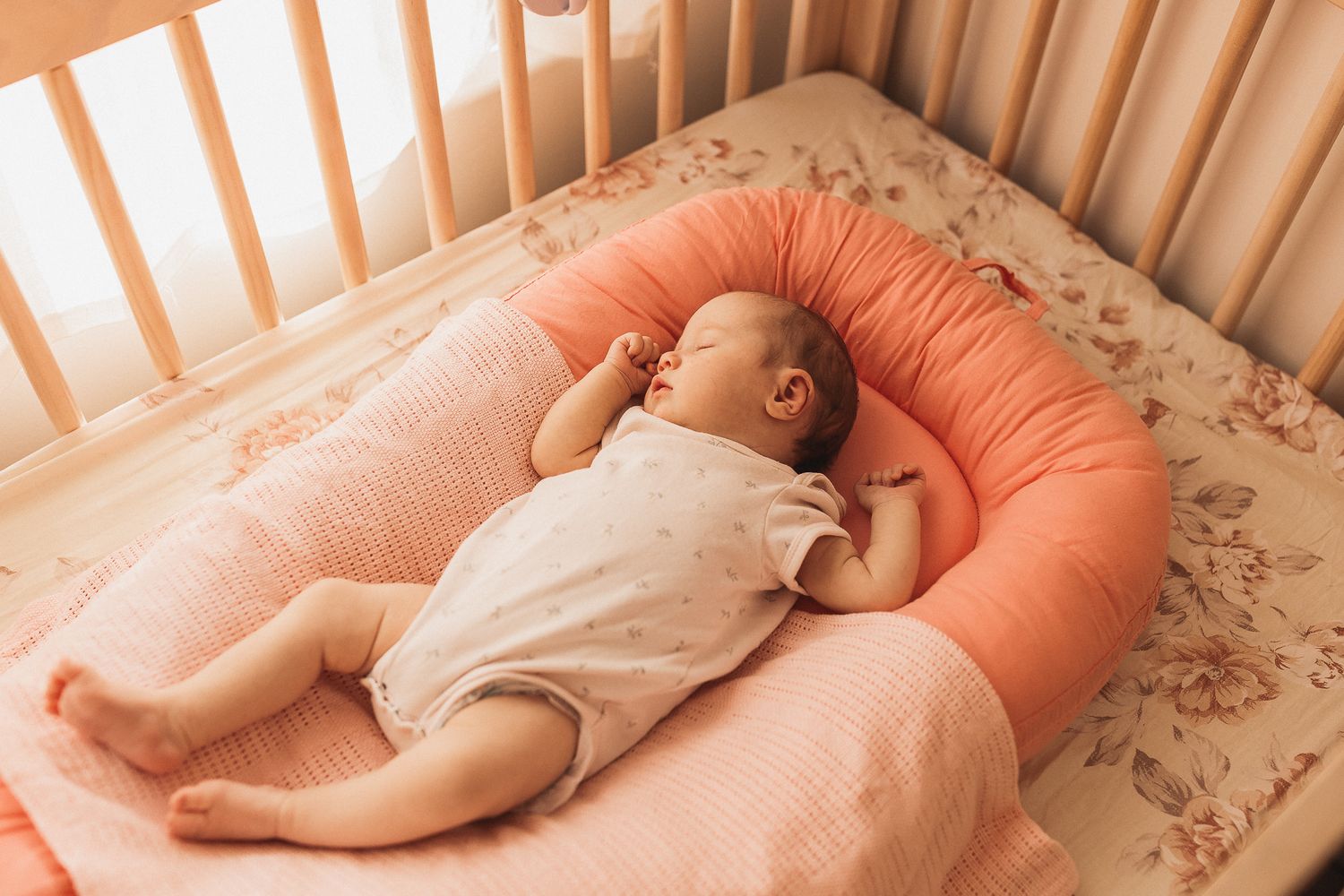A sleeping baby rests peacefully in a coral-colored lounger cushion inside a wooden crib.