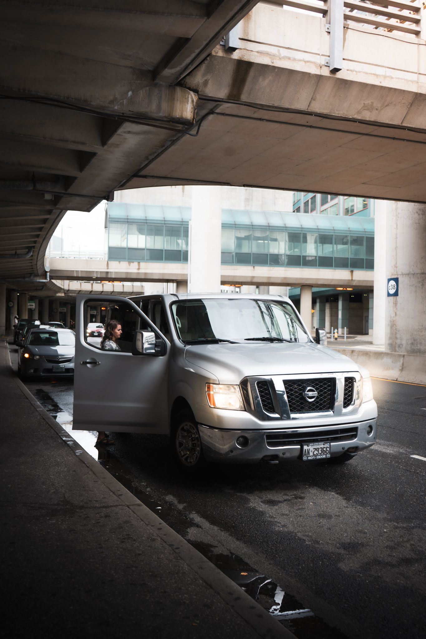 A matte gray Nissan NV cargo van parked under a concrete overpass with modern buildings visible in the background.