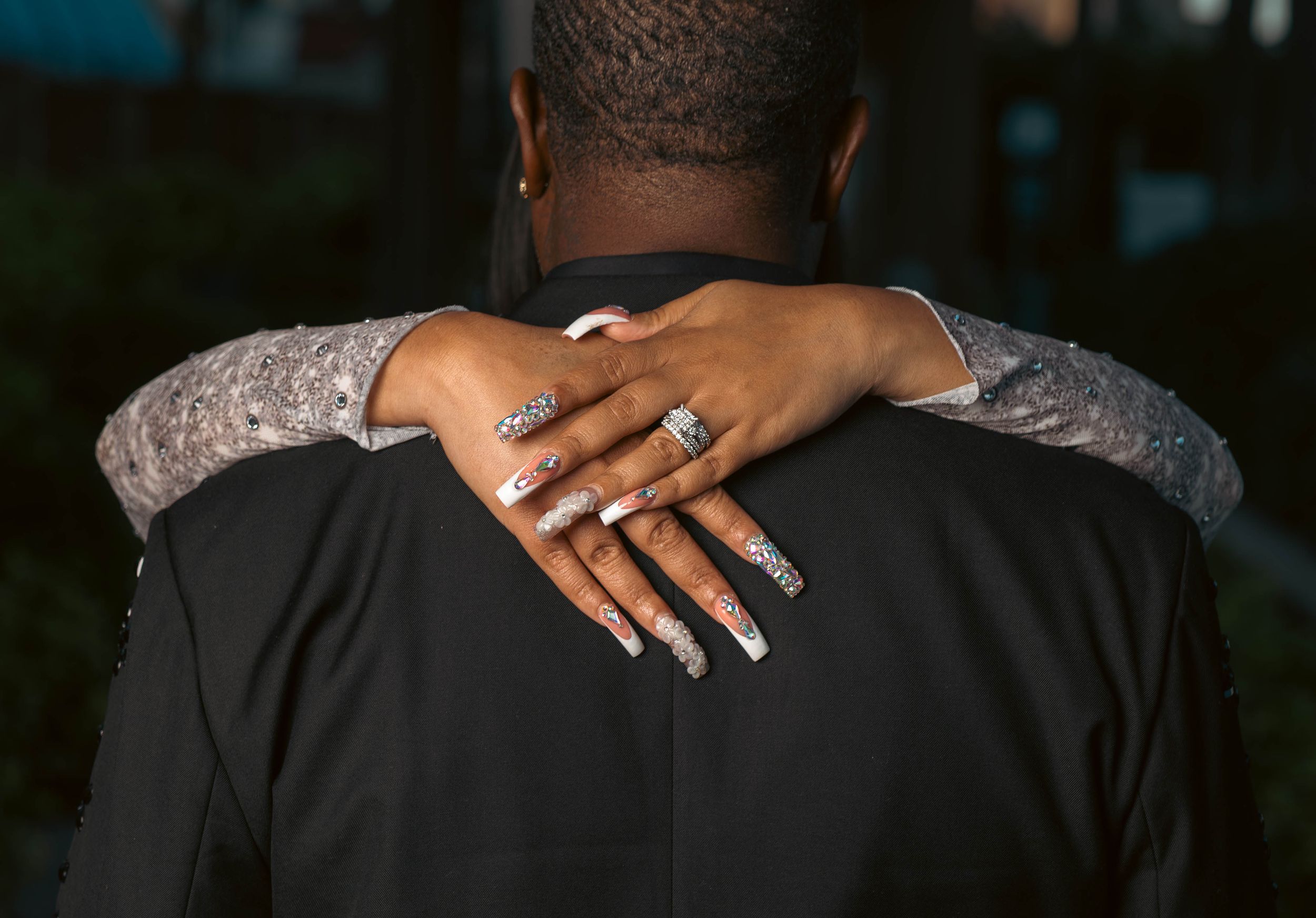 Close-up of a bride's diamond ring and manicured nails resting on groom's suited shoulder.