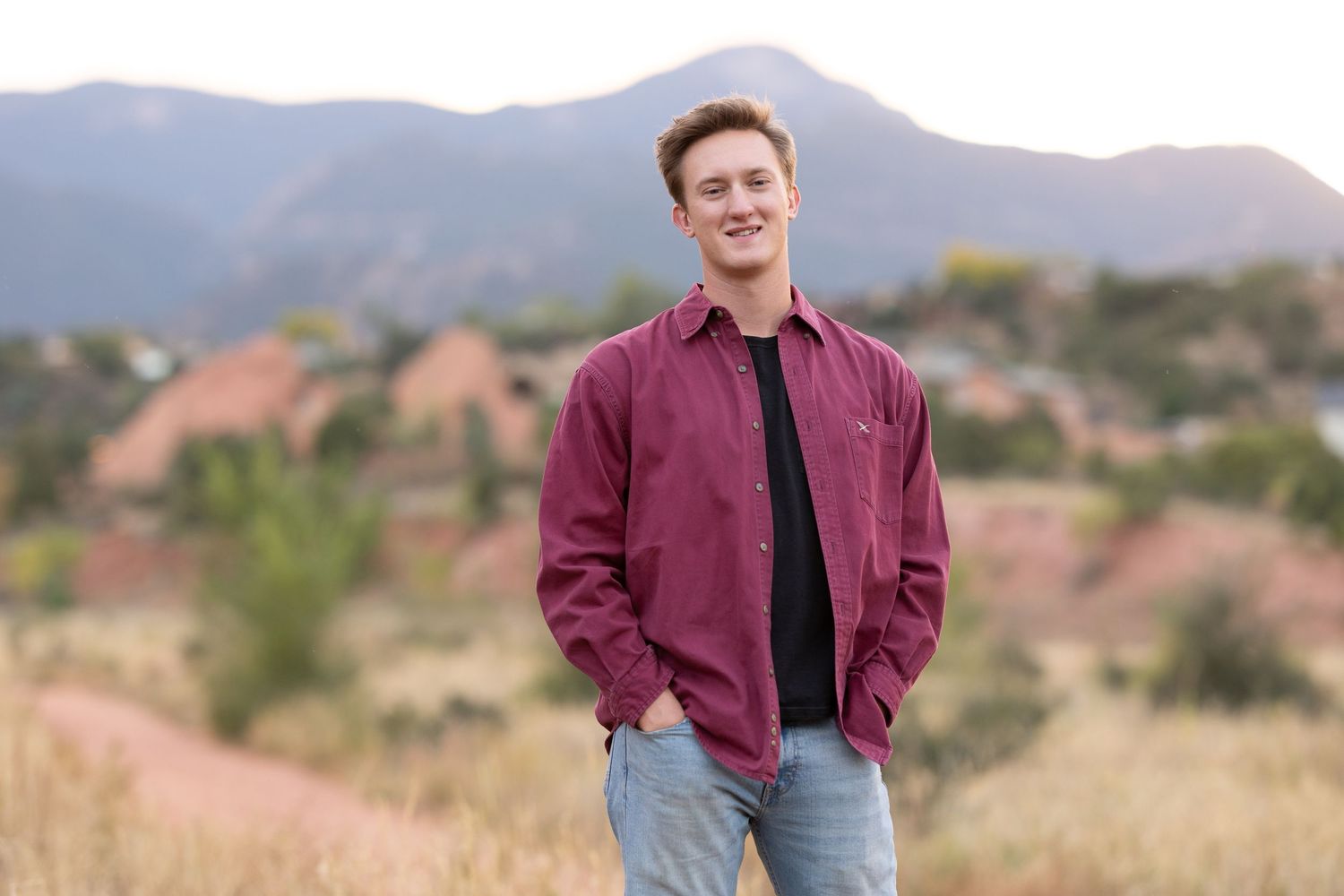 A person in a burgundy shirt stands in a field with mountains in the background.