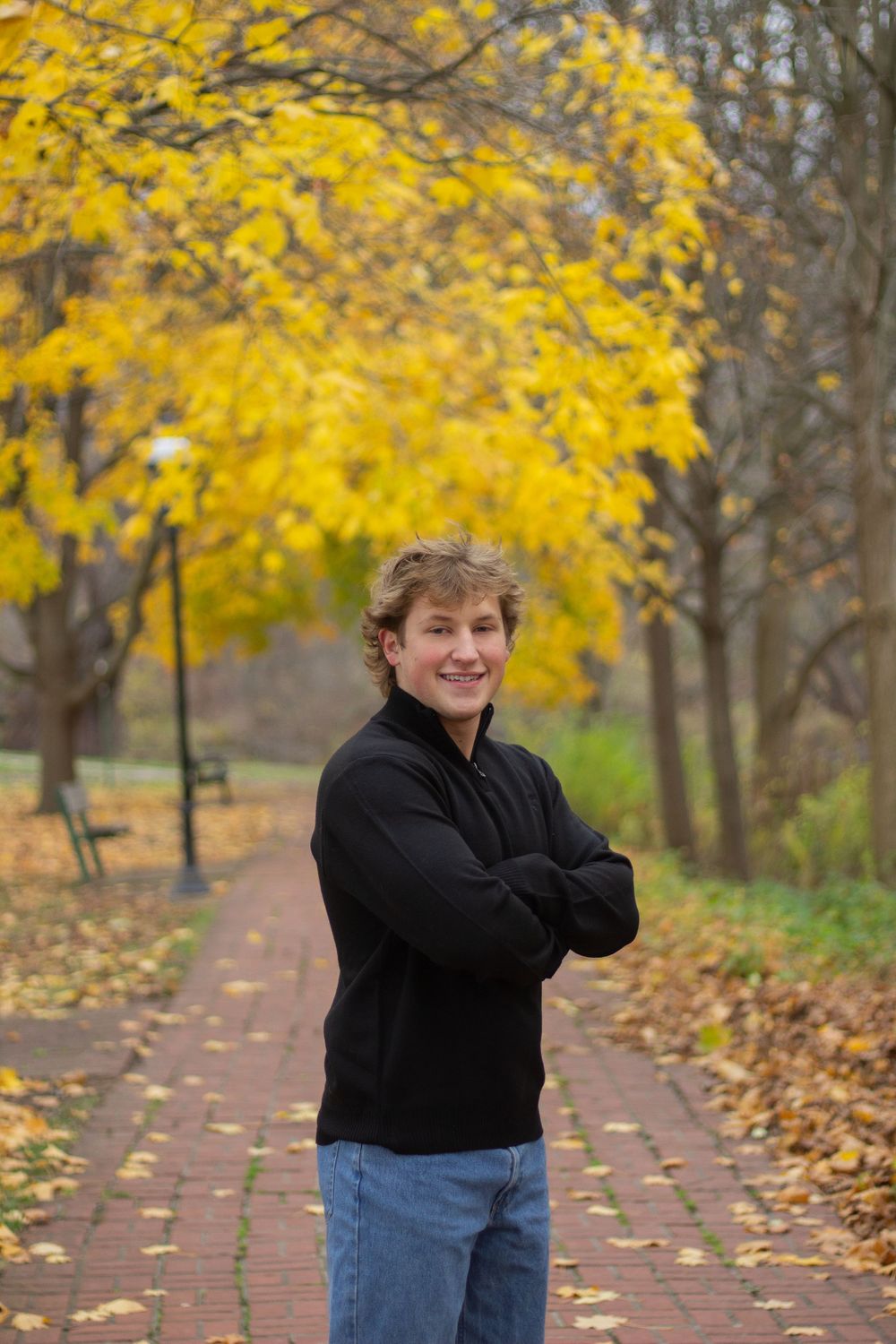 A person in a black sweater stands smiling on a brick path with vibrant yellow autumn foliage overhead during senior photos.