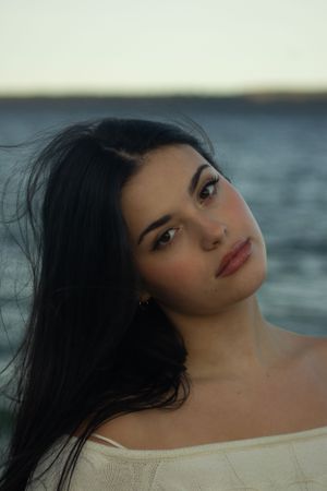 A person with dark hair poses against a moody ocean backdrop at sunset in an artistic portrait series during senior photos in Holland, MI.