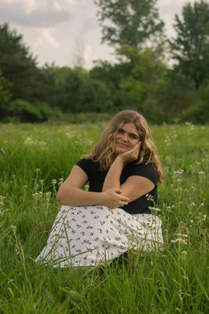 A girl wearing a black t shirt and a polka dot skirt, sits in a grassy flower field during her senior photo session in Flushing, Michigan.