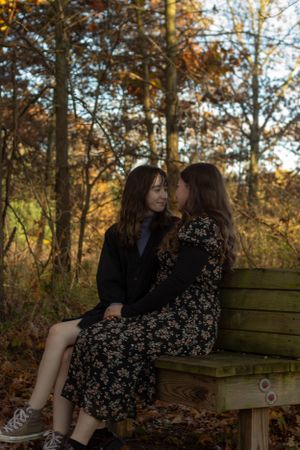 An LGBTQ couple sits together on a wooden bench surrounded by autumn trees with golden leaves in a park setting during their engagement.