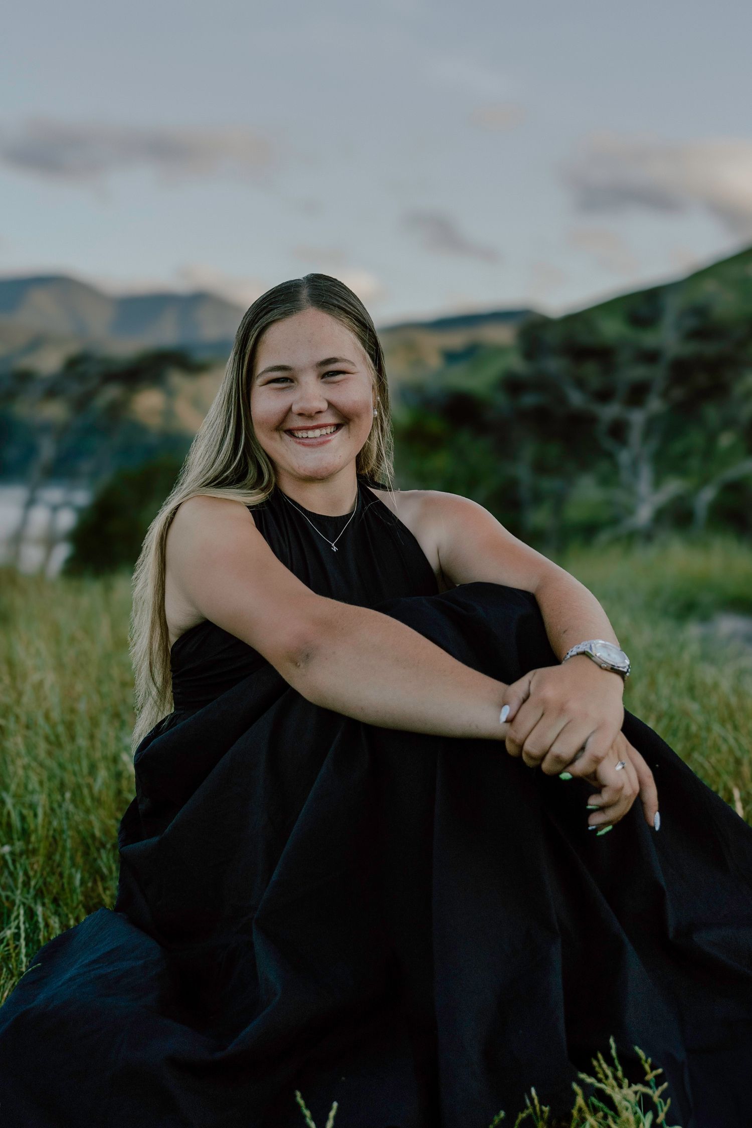 Person in black dress sits in grassy field with mountains and water in background.