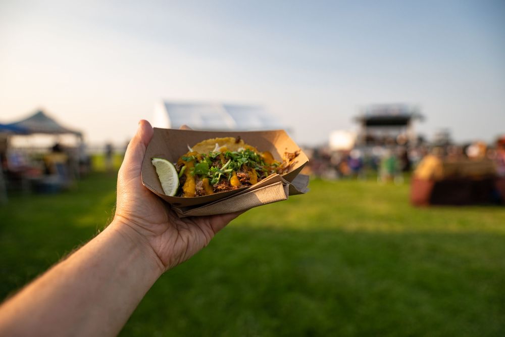 A hand holds a tacos with toppings against a blurred outdoor landscape backdrop.