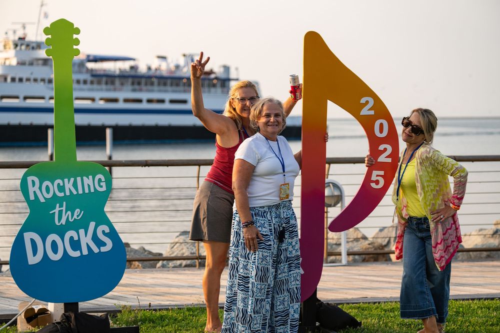 Group of tourists stand along a waterfront plaza near cruise ships with colorful bird-shaped signage at Waterfront Park.