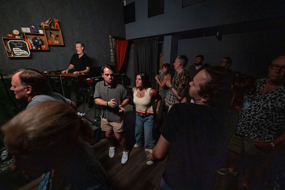 A dimly lit crowd gathers in a small venue space watching a performance in a dark room.