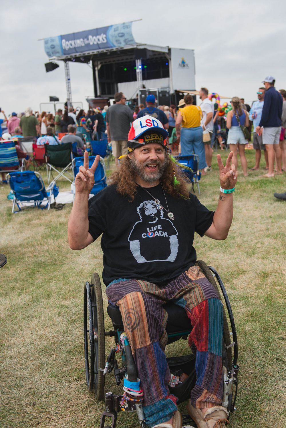 Festival goer in wheelchair wearing USA hat and colorful pants gives peace signs at outdoor music event.