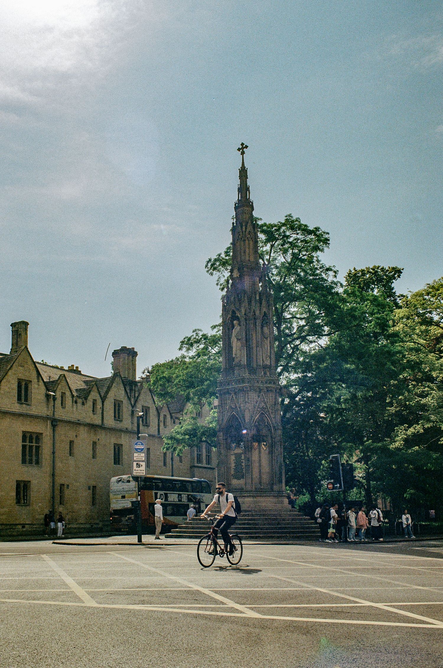 Victorian Gothic memorial spire in a town square with historic buildings and cyclists.