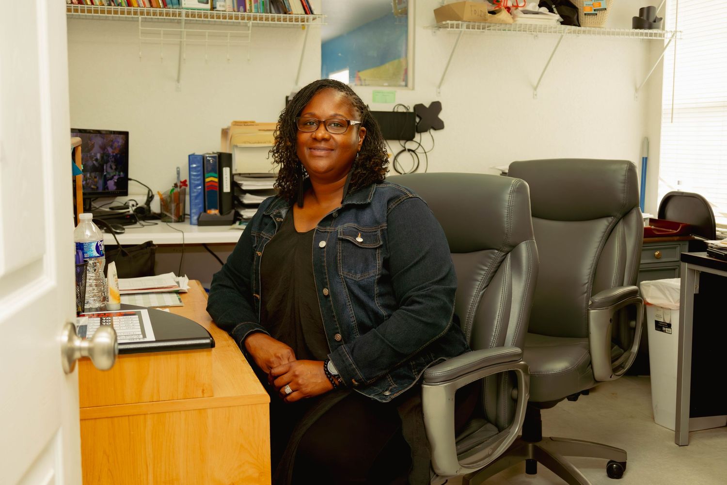 Black woman smiling while leaning on office desk with bookshelves in background.