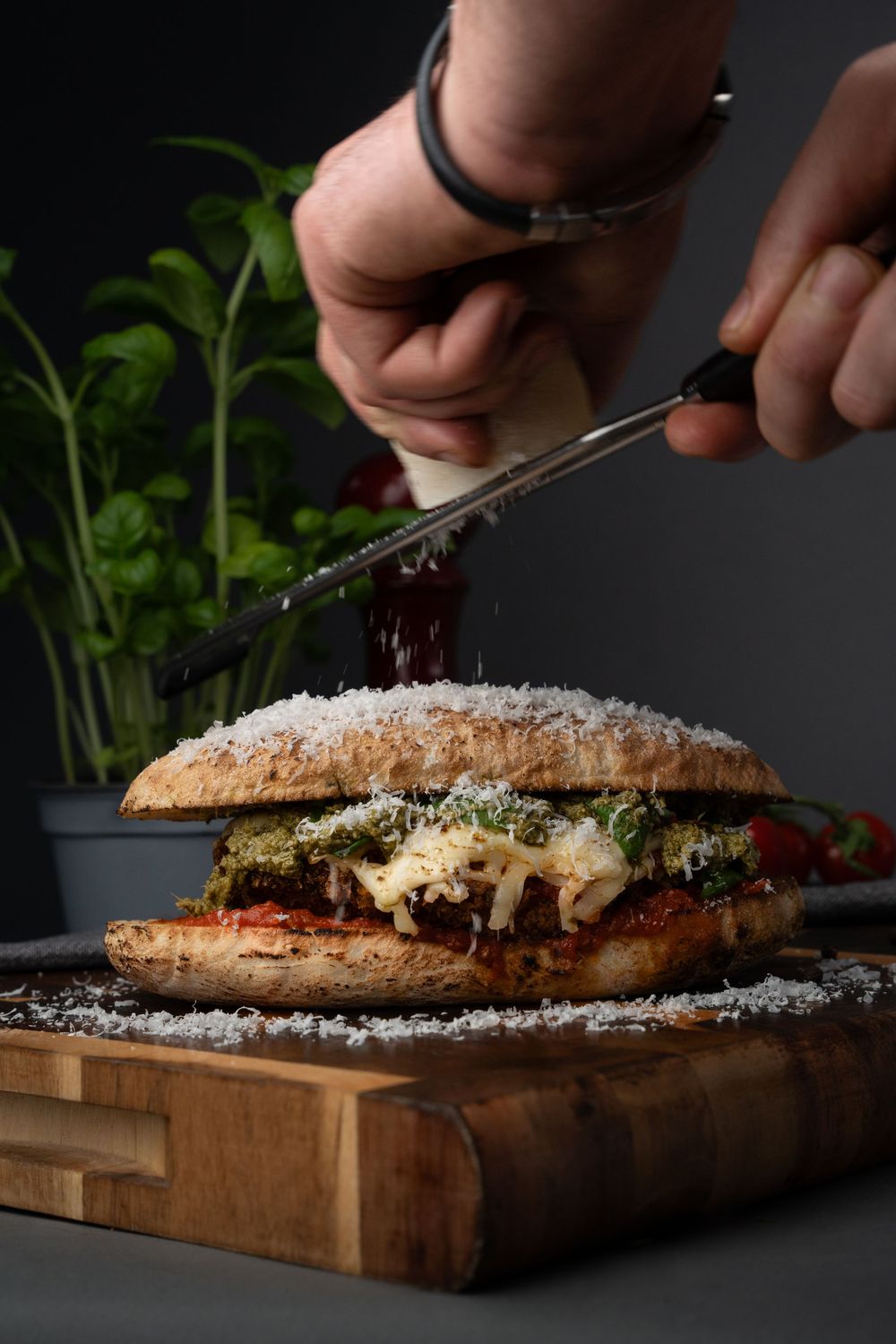 Hands sprinkling grated cheese over a rustic sandwich on wooden cutting board in moody lighting.