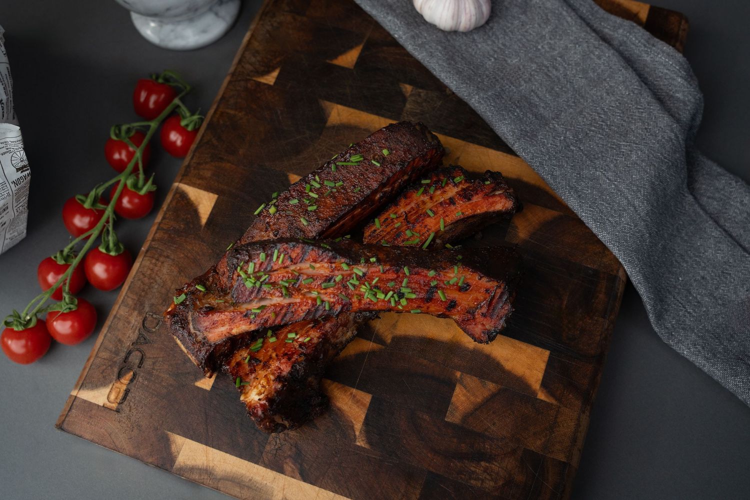 Grilled beef ribs with tomatoes on a dark wooden cutting board with dramatic lighting.