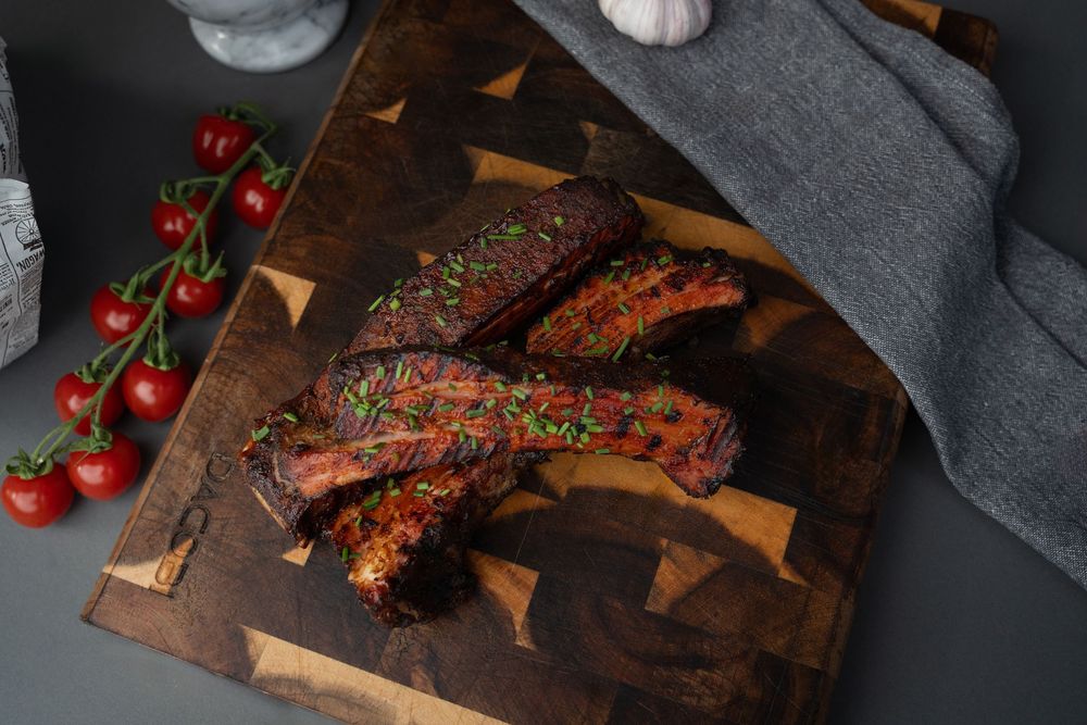 Grilled beef ribs with tomatoes on a dark wooden cutting board with dramatic lighting.
