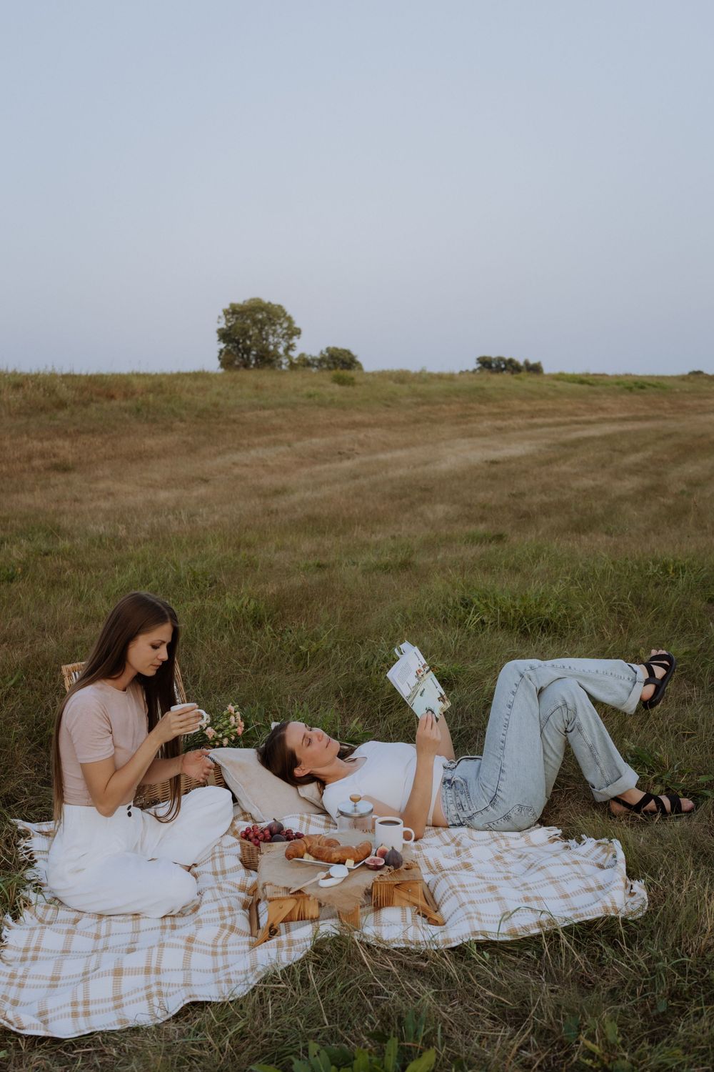 Two friends relaxing on picnic blanket while reading and enjoying snacks in grassy field.