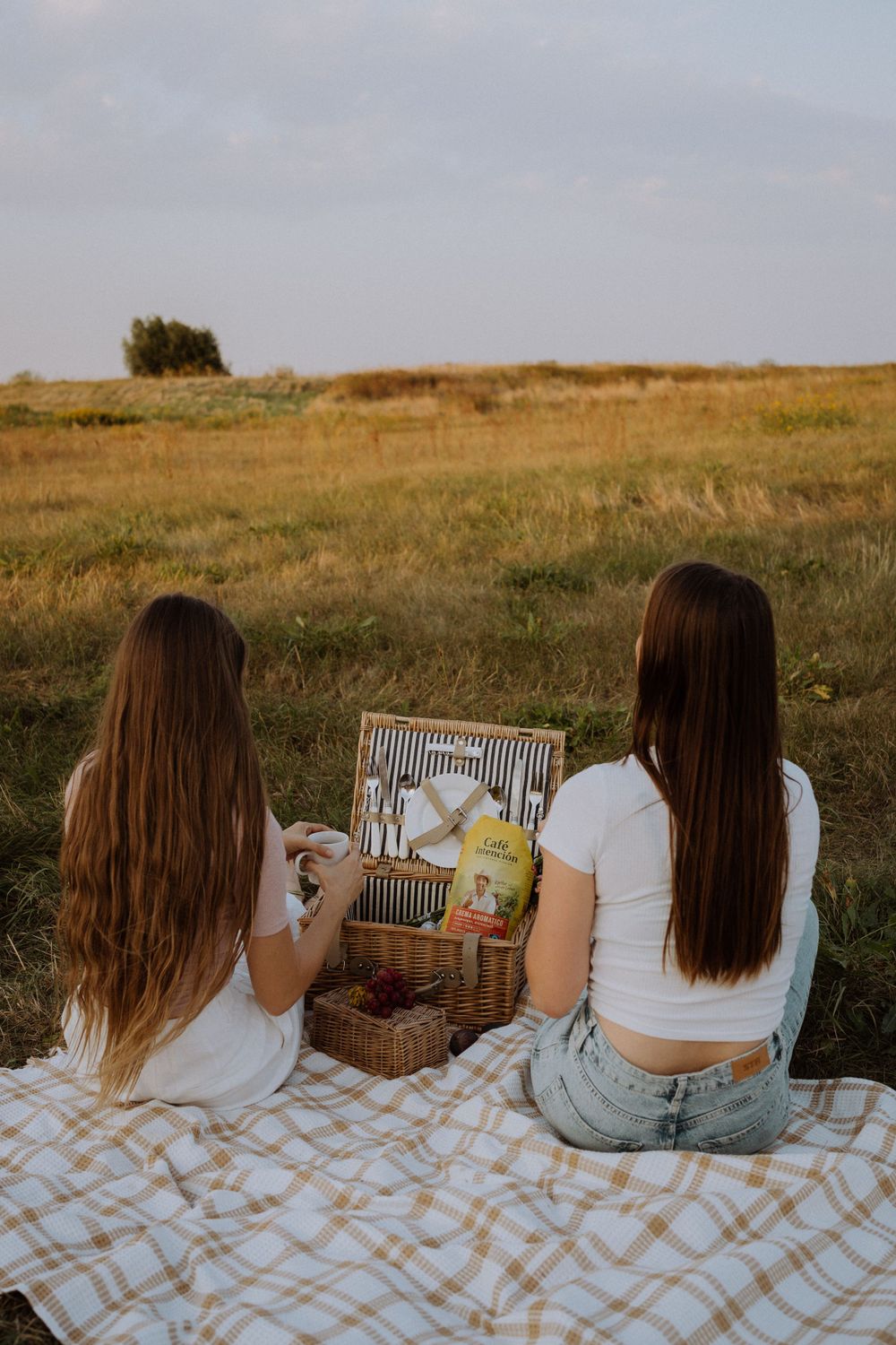 Two friends enjoying a picnic on a blanket while looking through a wicker basket at sunset.