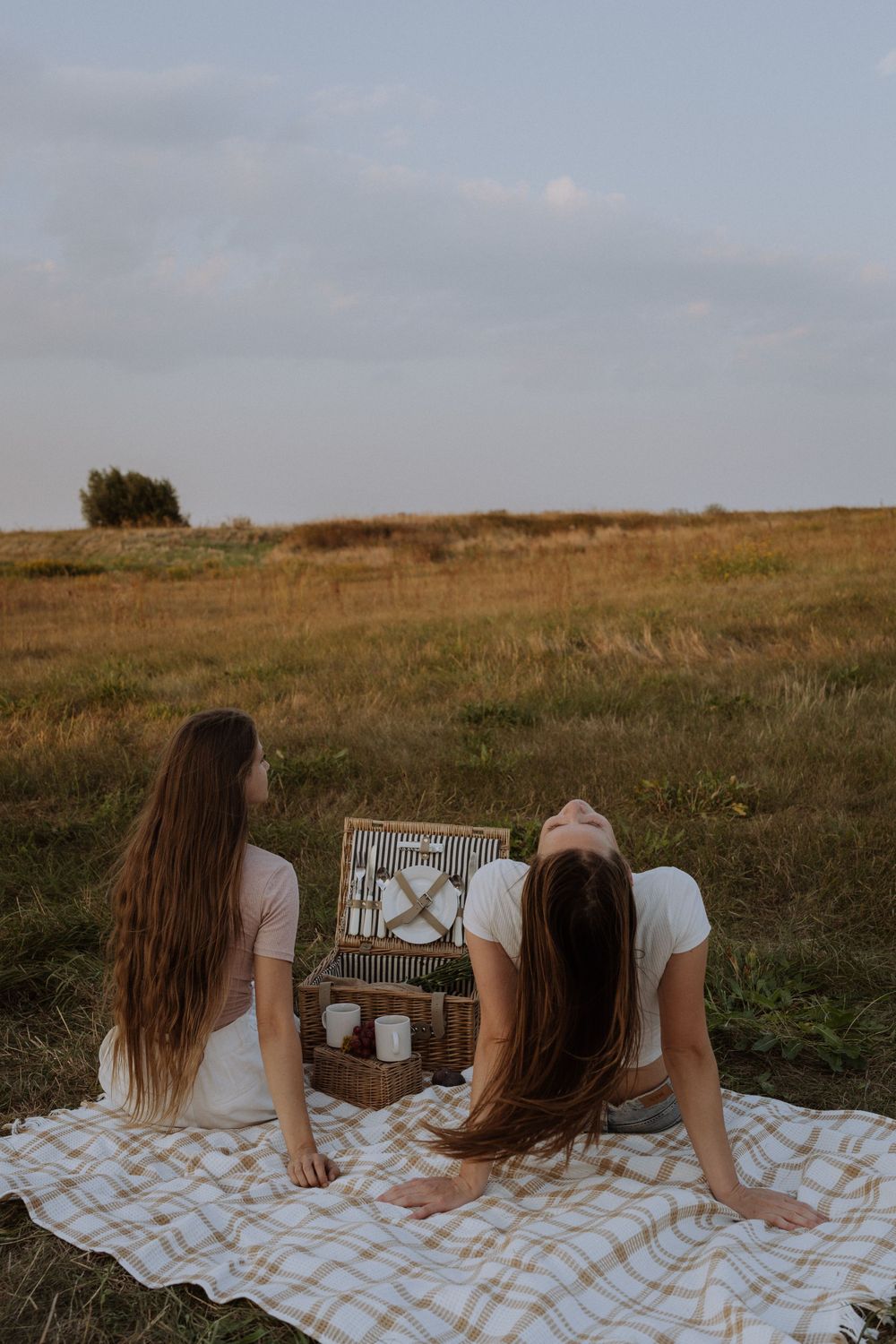Two friends lounging on plaid blanket beside picnic basket at golden hour.