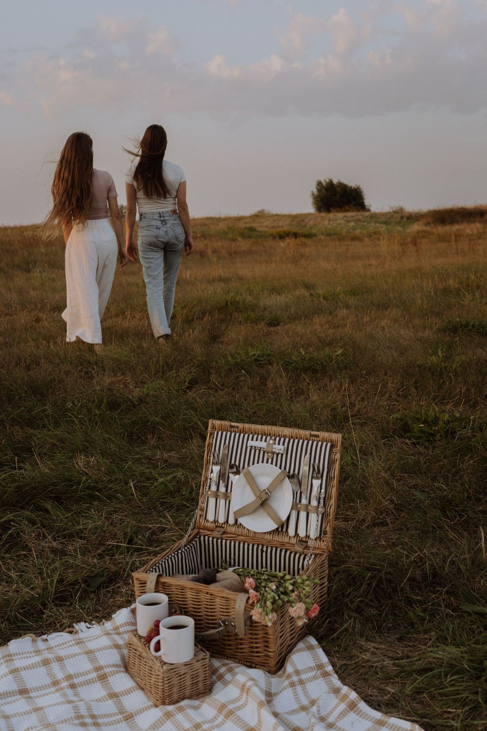 Two friends walking in a field at sunset with a picnic basket and blanket set up in the foreground.