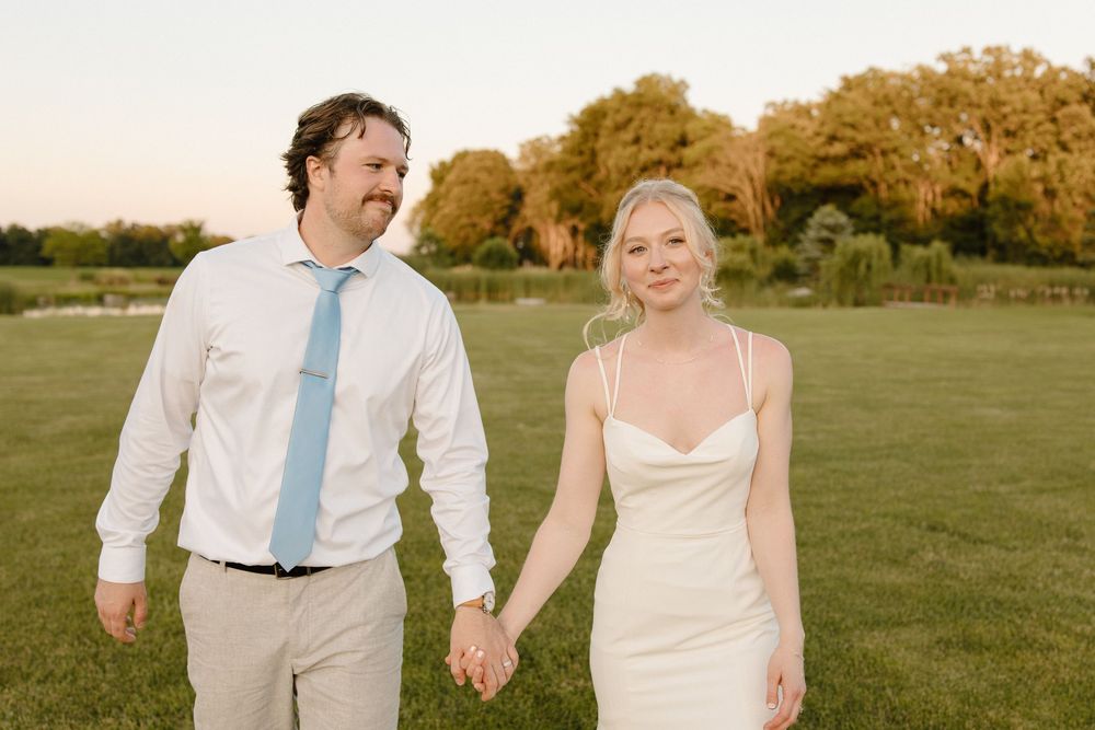 A newlywed couple holds hands while walking together on a grassy field during golden hour.