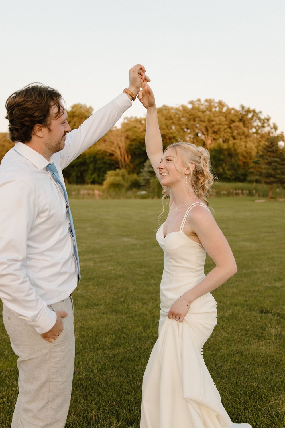 A couple dances together on a grassy field at sunset wearing elegant white wedding attire.