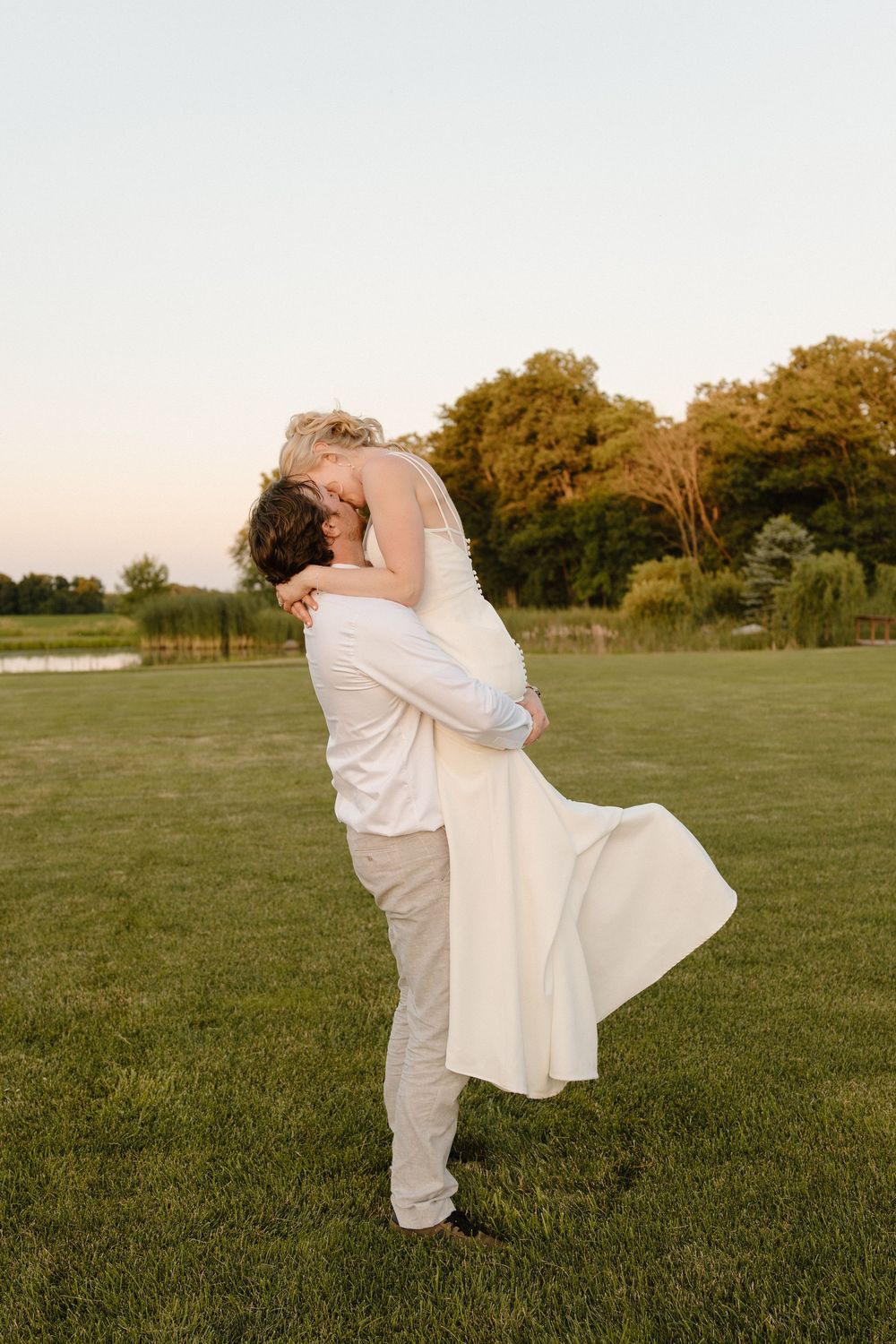A couple in white outfits embrace in a romantic spinning dance pose on a grassy field at sunset.
