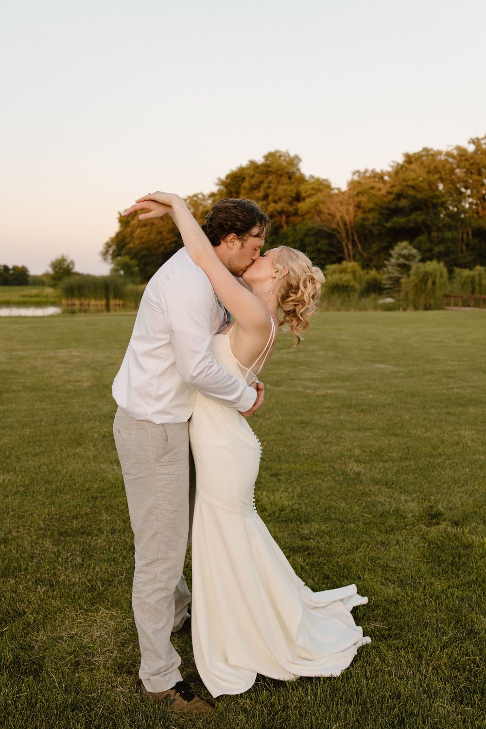 A romantic wedding couple shares an intimate kiss in a golden-lit field during sunset, her white dress flowing in the breeze.