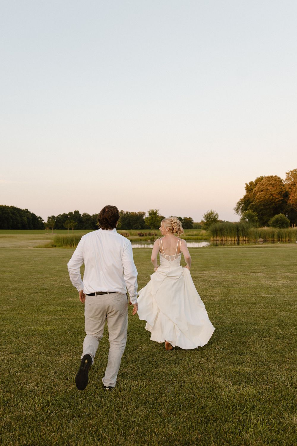 A couple in wedding attire walks across a green lawn at sunset with trees in the background.