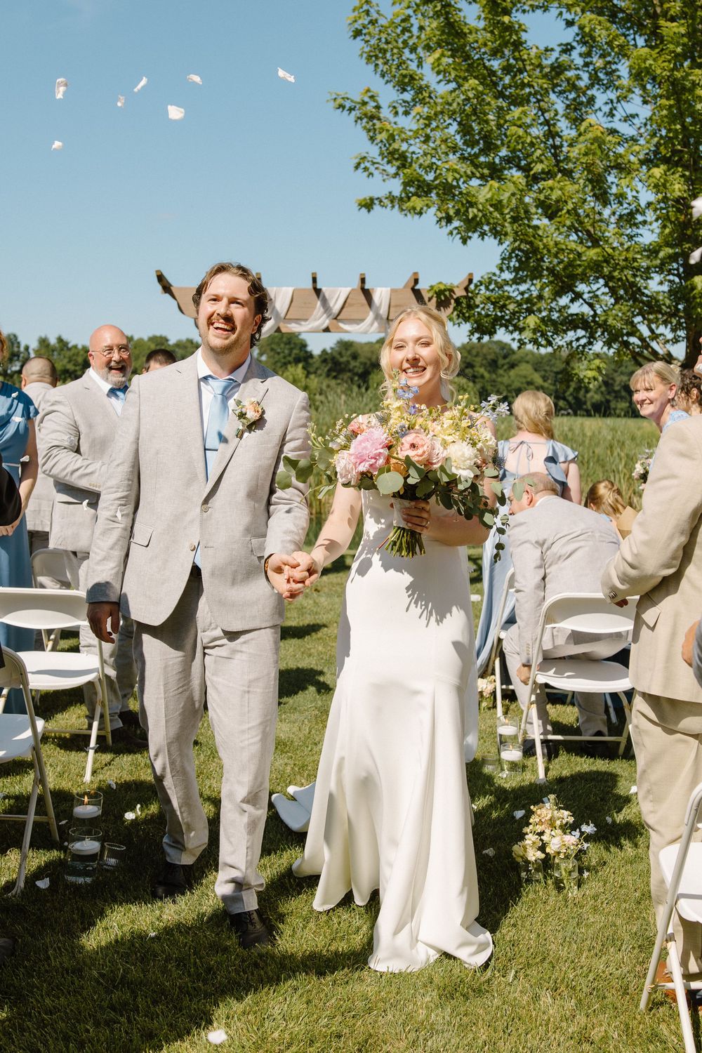 A joyful couple walk down an outdoor aisle on a sunny day with white chairs and confetti floating in the air.
