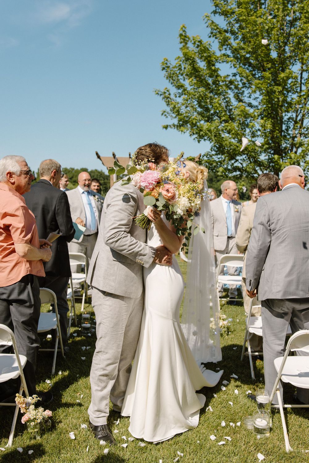 Wedding guests celebrate at an outdoor garden ceremony with flower petals in the air and white chairs arranged in rows.