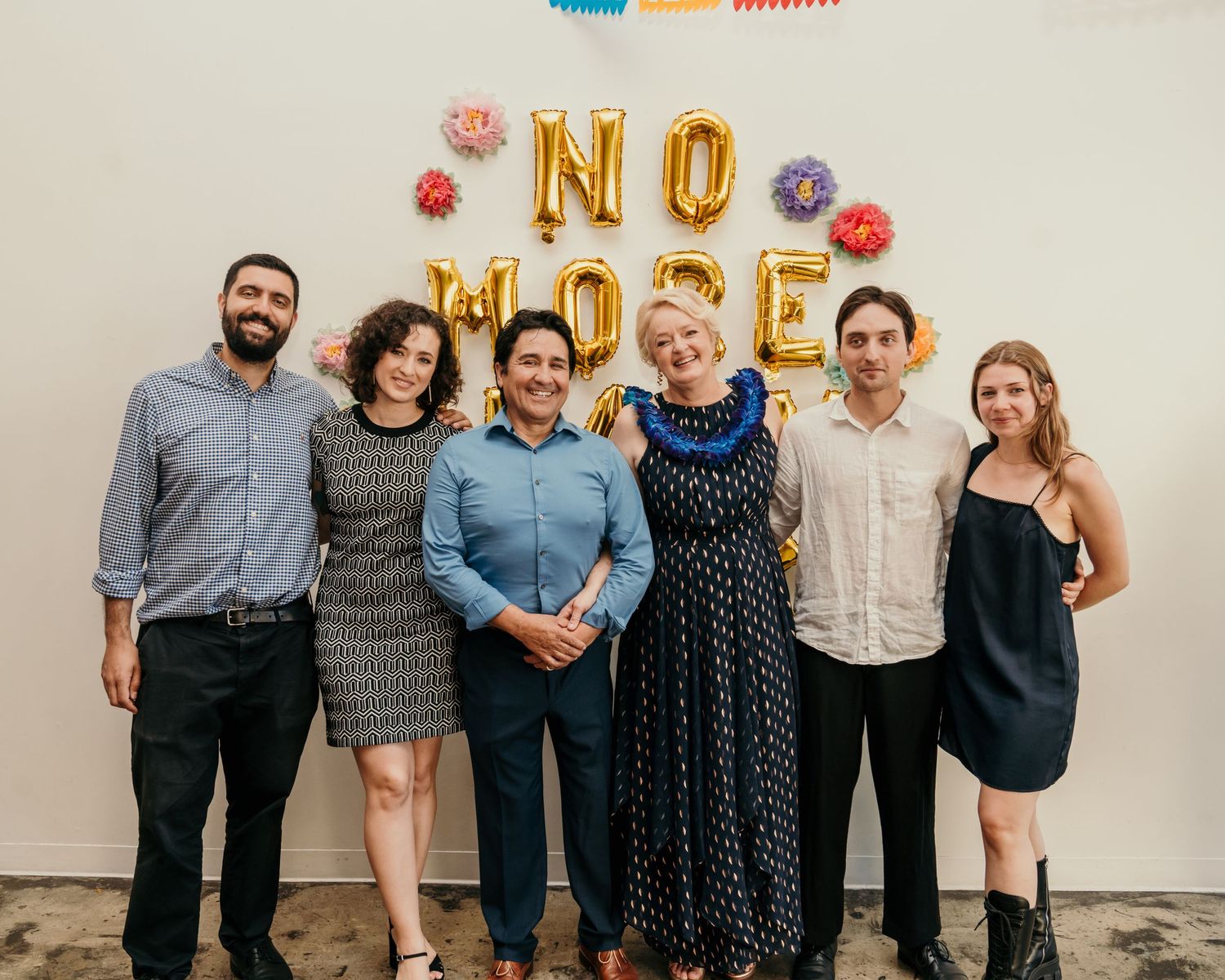 A group celebrating in front of gold NO MORE balloon letters with colorful decorative elements.