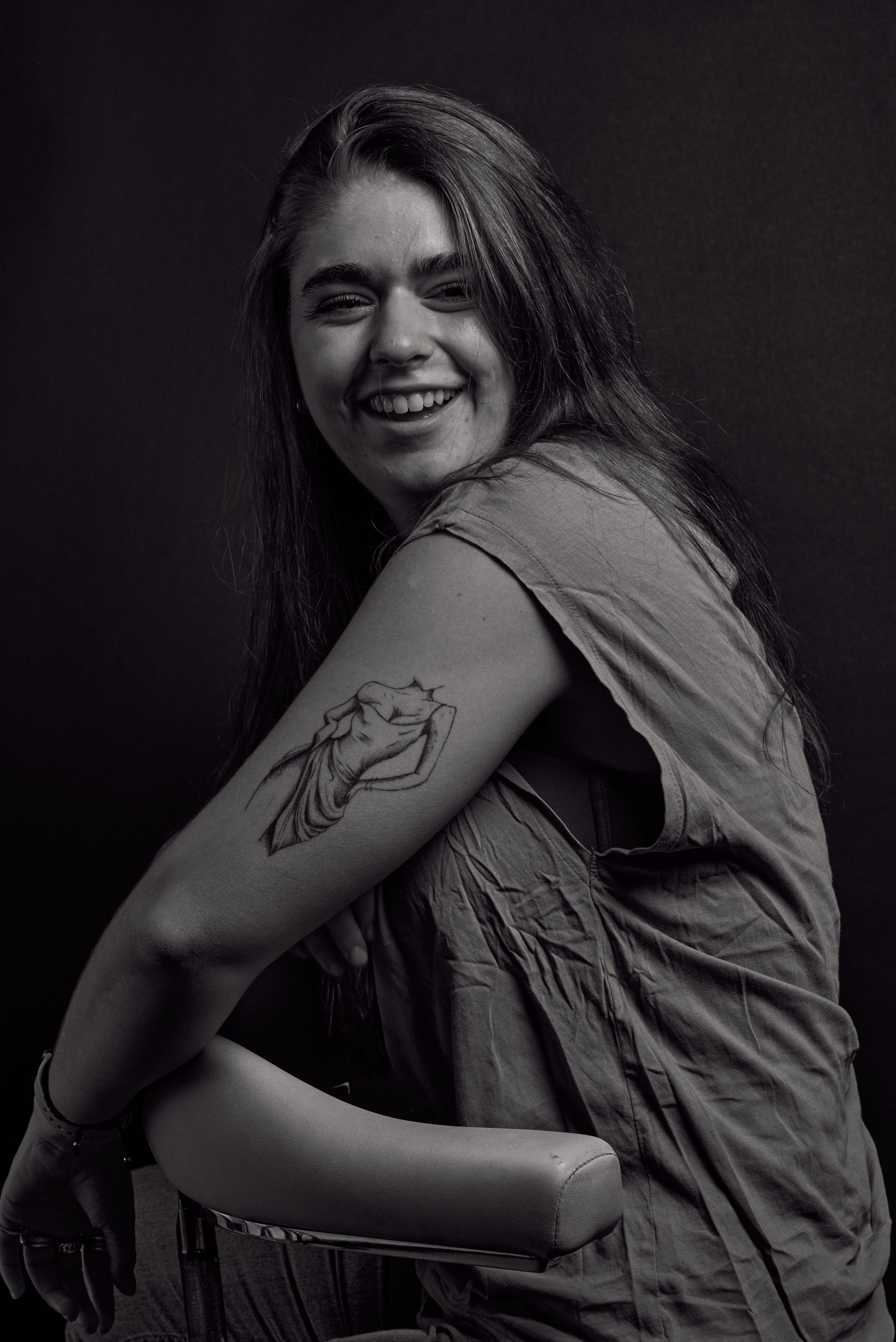 Smiling young woman with long hair reveals a tattoo on her arm in black and white photo.
