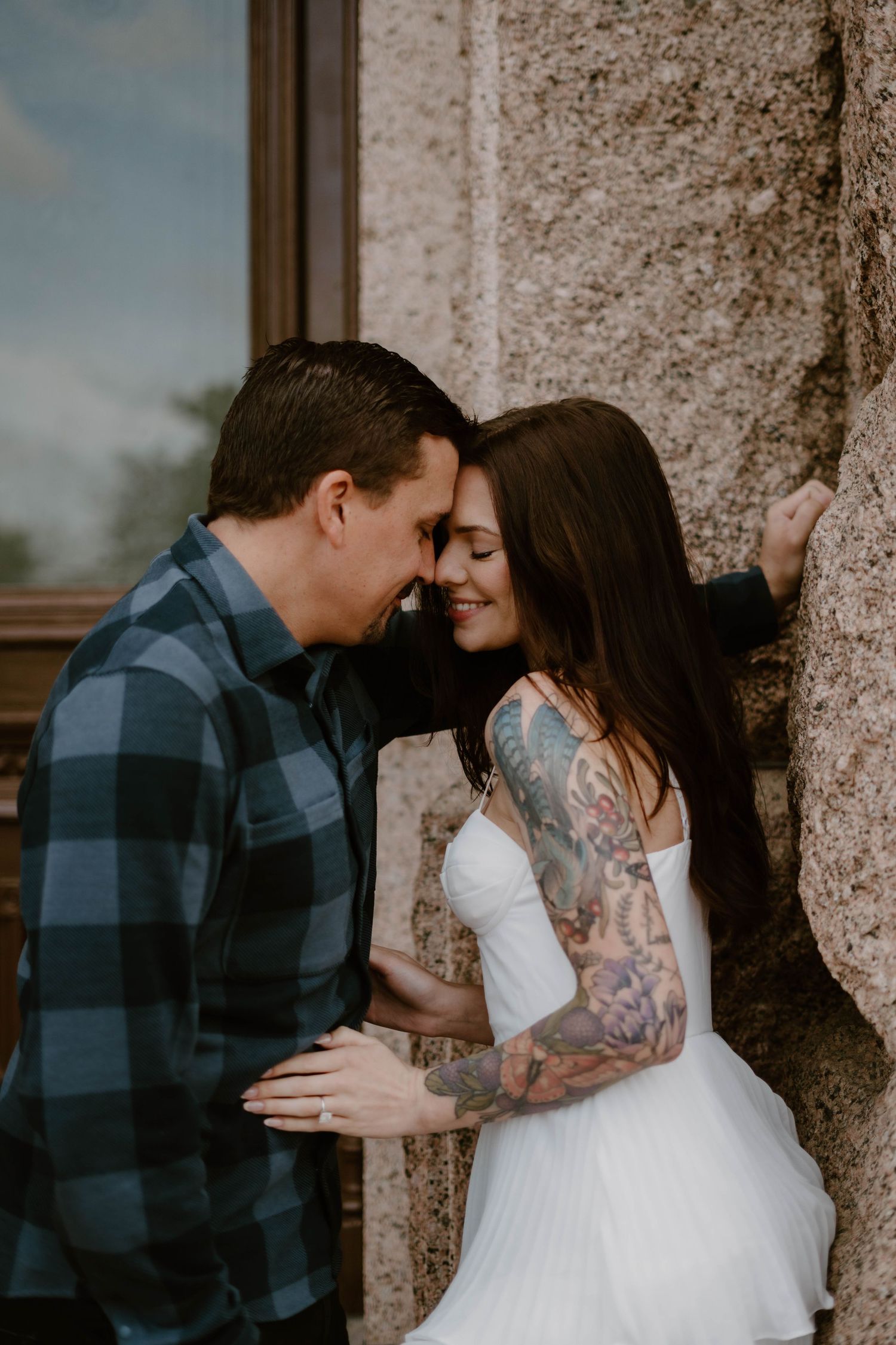 A romantic couple shares an intimate moment against a stone wall, with the bride wearing a white dress and showing tattoo sleeves.