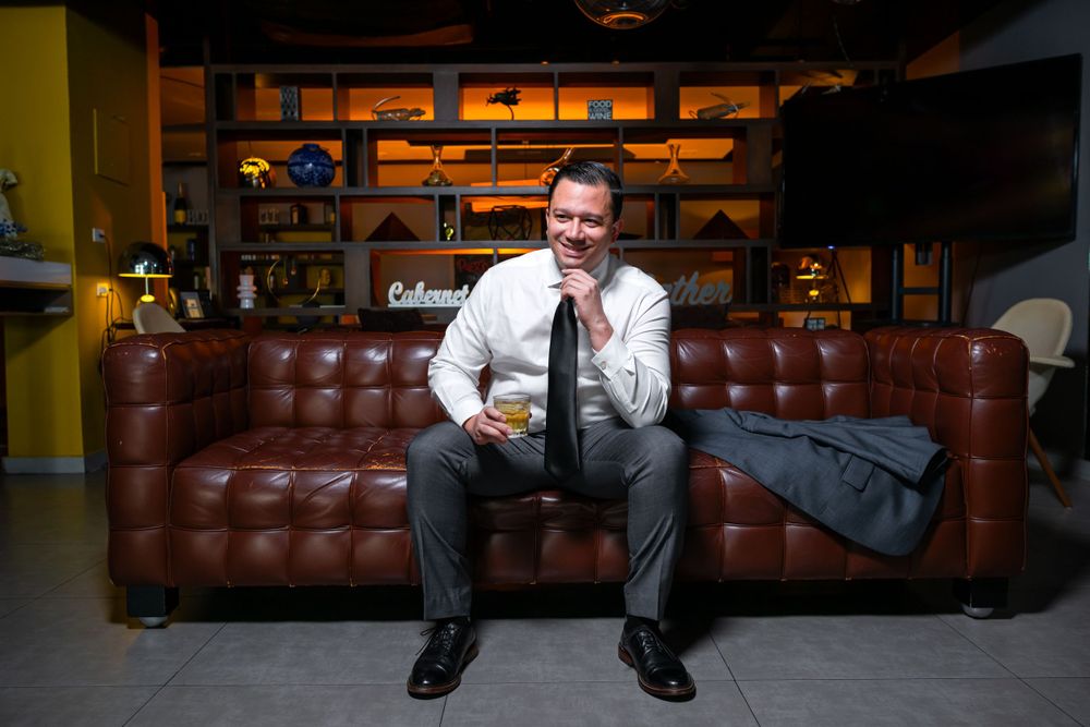 Man in formal attire sits casually on brown leather couch in modern lounge area.