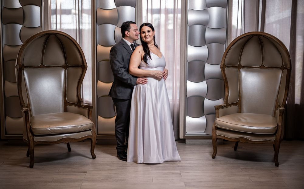 A couple poses between two high-backed chairs in an elegant indoor setting.