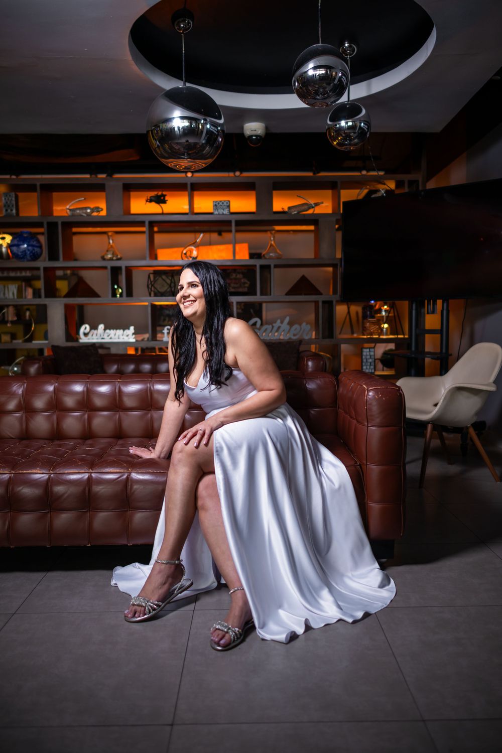Woman in elegant white gown sits on brown leather couch in modern lounge setting.