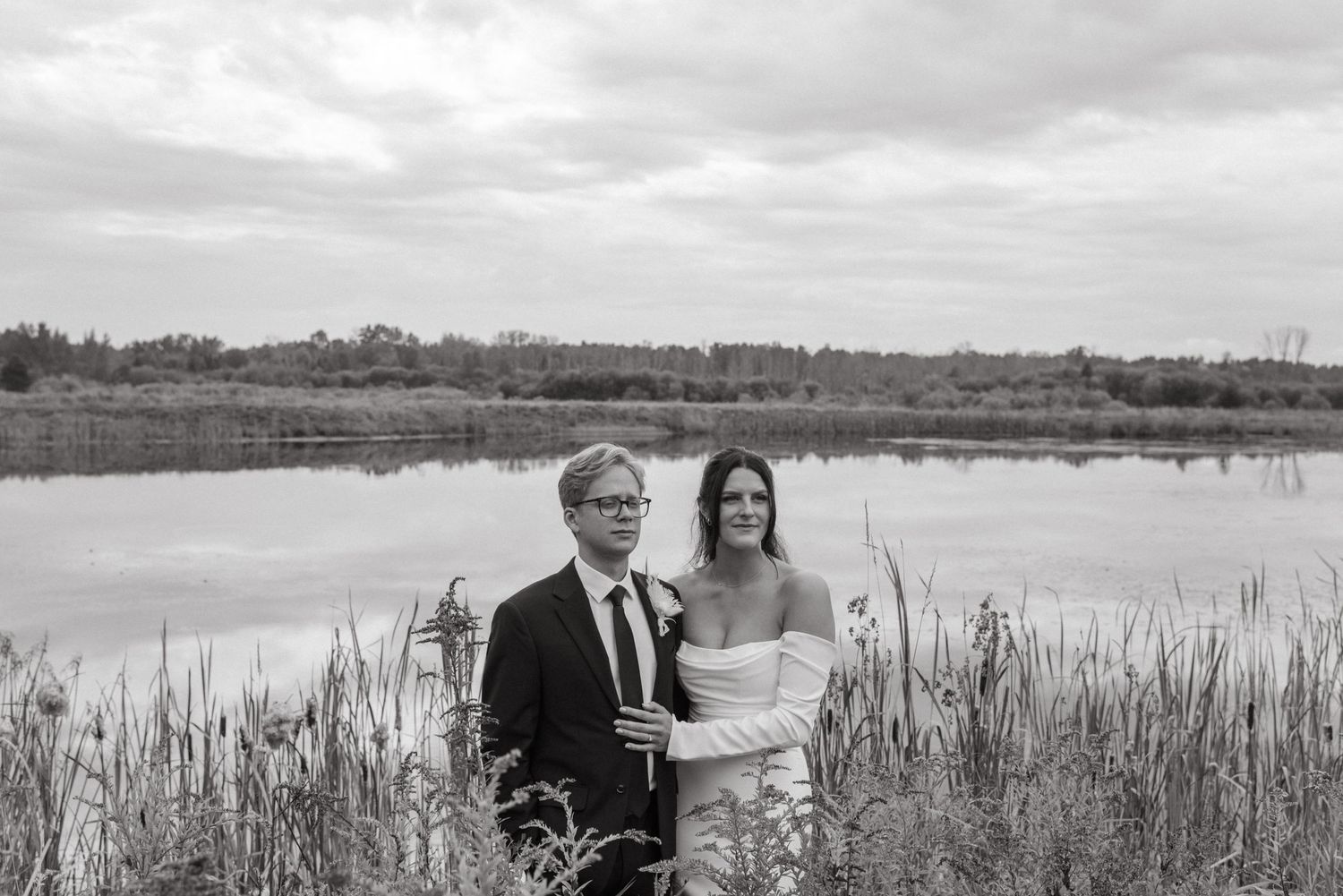 Couple sharing a romantic moment in tall grass by a serene lake, bride in white gown and groom in black suit.