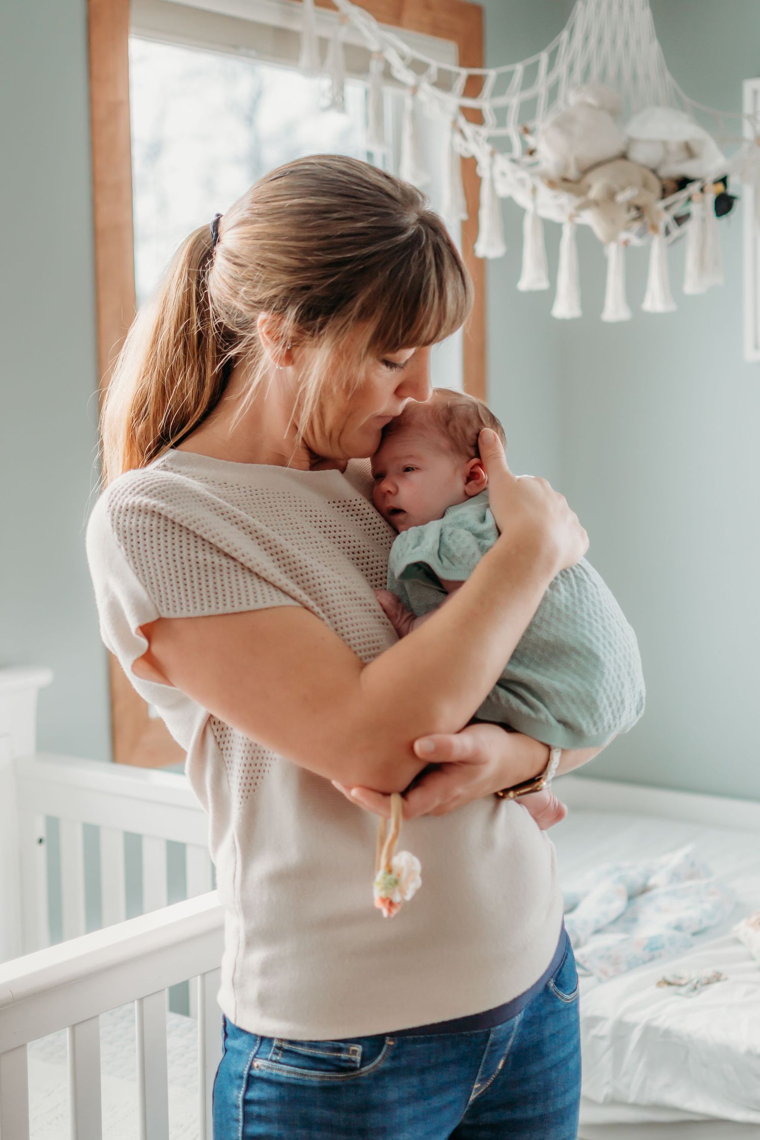 A mother tenderly kisses her newborn baby while standing in a softly decorated nursery room.