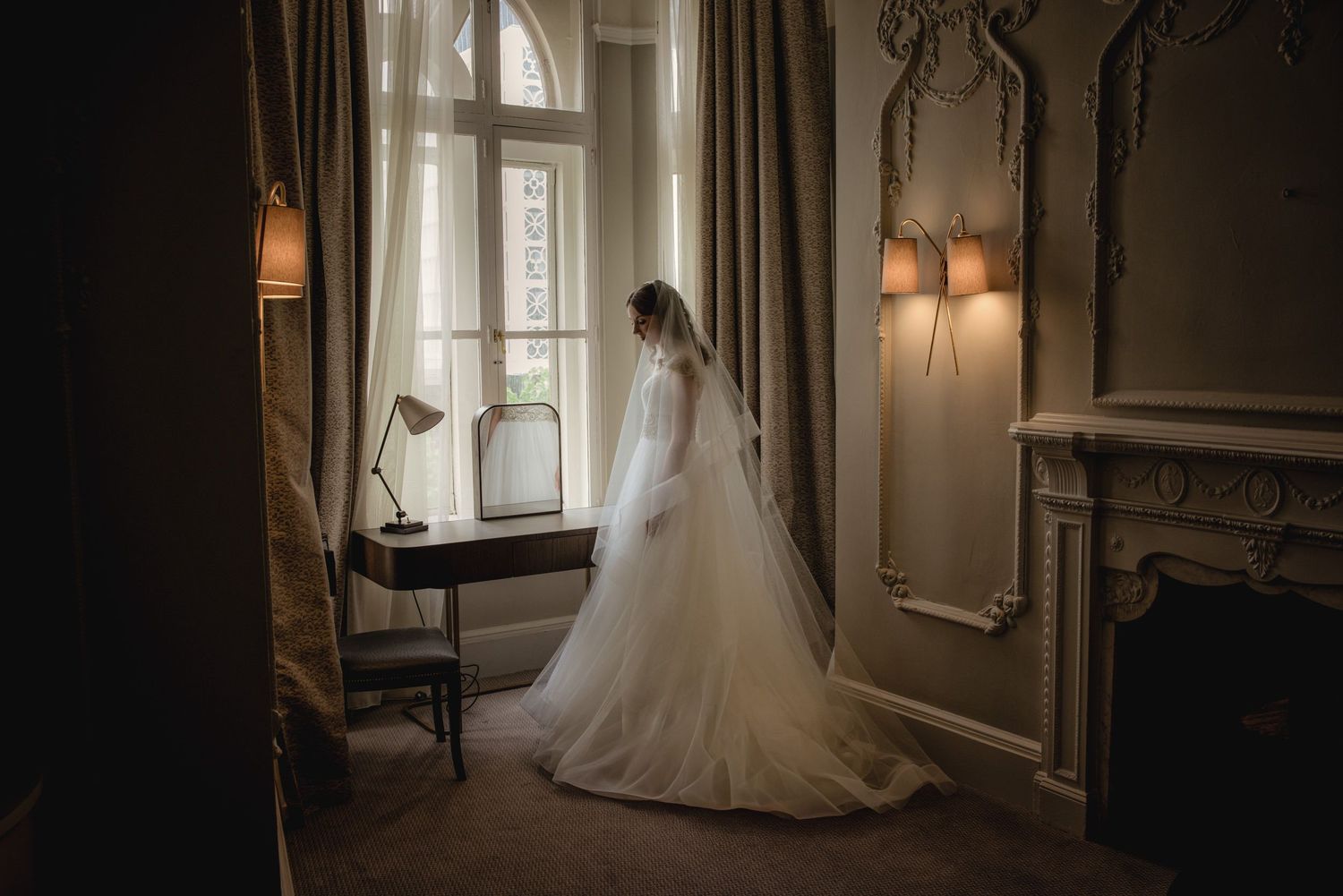 Bride in elegant white wedding gown with long veil standing by a vintage window in a classic ornate room.
