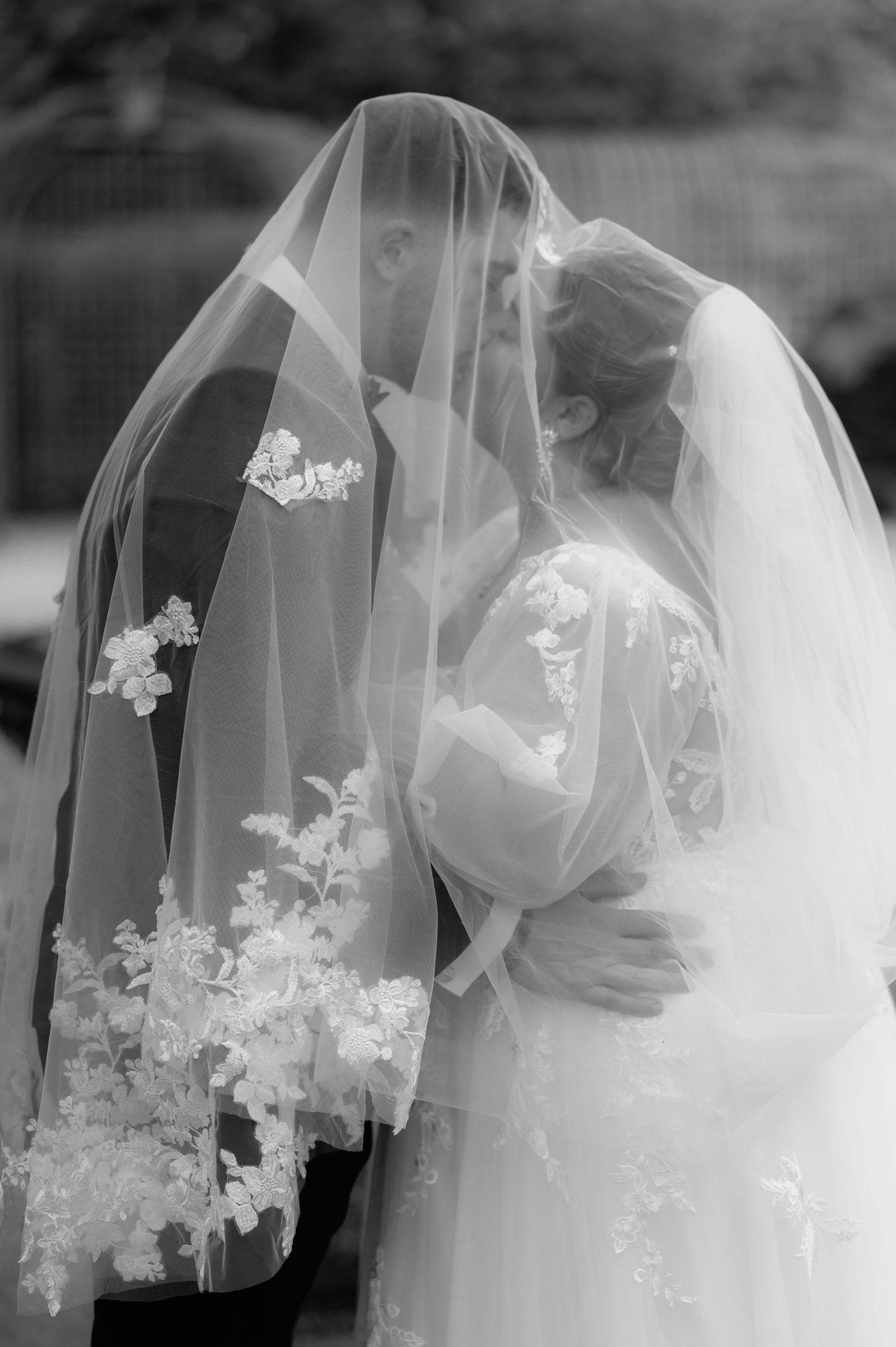 Photo en noir et blanc de la mariée et du marié s'embrassant sous un voile de mariage en dentelle florale.