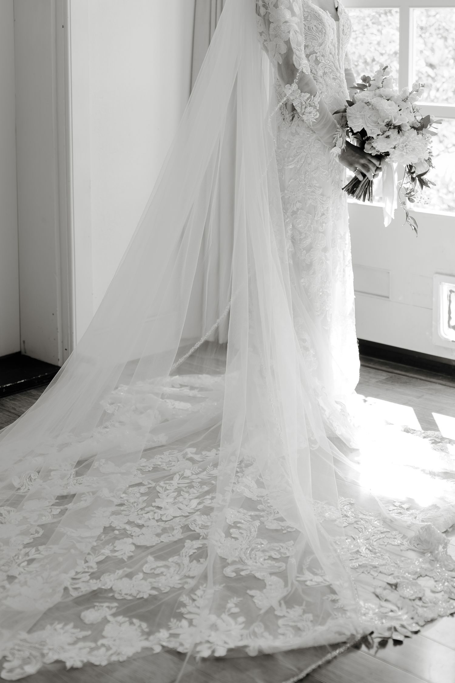 Bride in lace wedding gown with long cathedral veil holding white bouquet, standing by window in bright room.