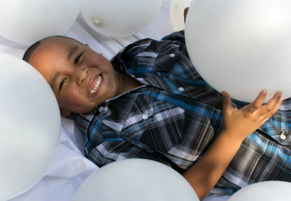 A young boy surrounded by white balloons smiles while holding a white balloon.