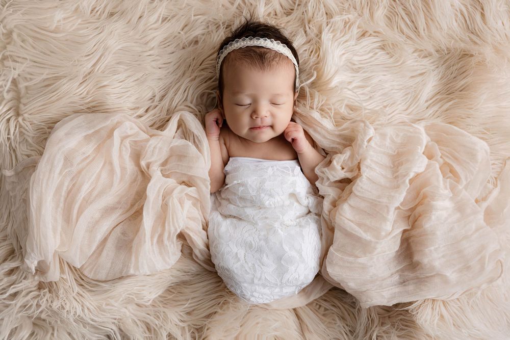 Sleeping newborn baby wrapped in white swaddle with headband on fluffy beige fur rug.