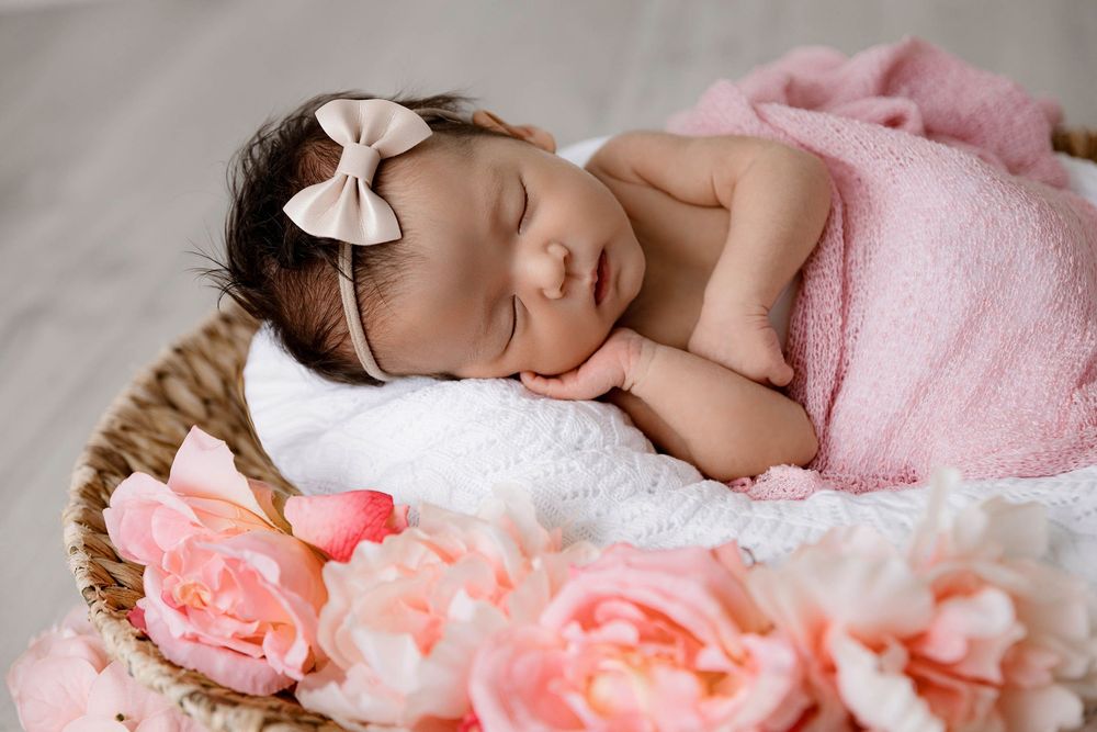Sleeping newborn baby girl in pink wrap with bow headband resting in wicker basket surrounded by pink roses.