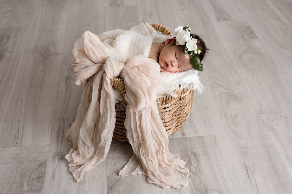Sleeping newborn baby in floral crown nestled in wicker basket with cream fabric bow on gray wood floor.