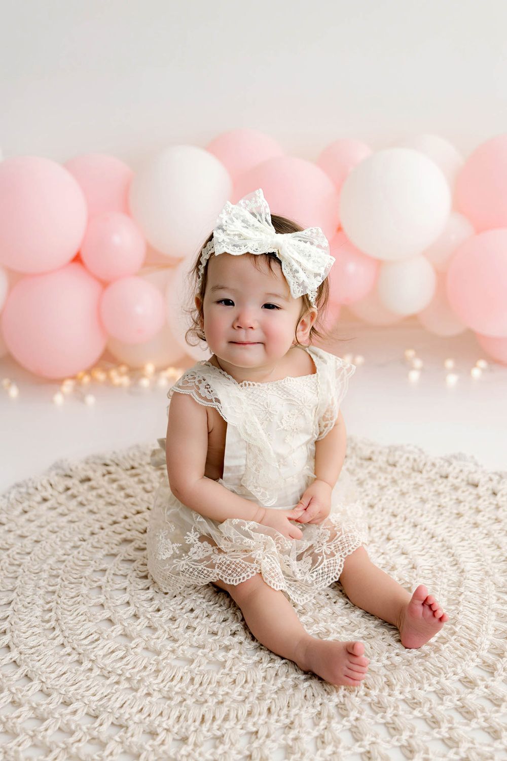 Smiling baby girl in white lace dress and bow headband sits on crochet rug with pink and white balloon backdrop.