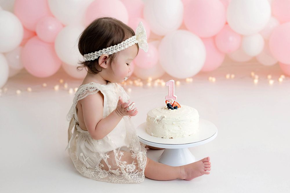 Baby girl in white lace dress reaches for first birthday cake with pink and white balloon backdrop.