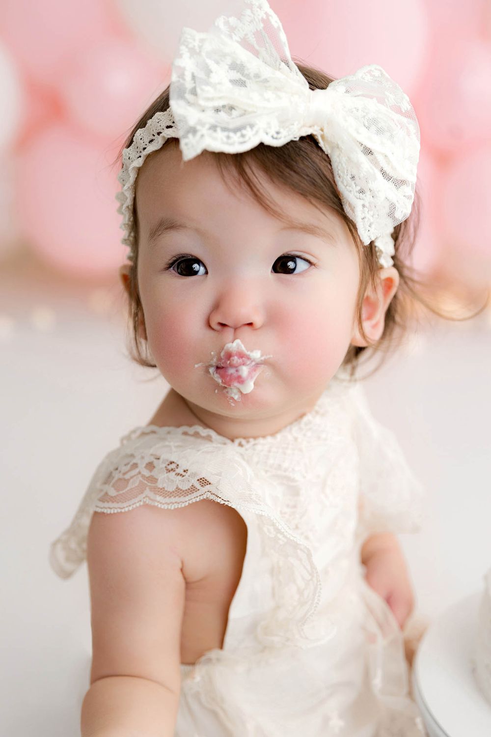 Close-up of baby girl in white lace dress and bow headband with birthday cake frosting on her lips.