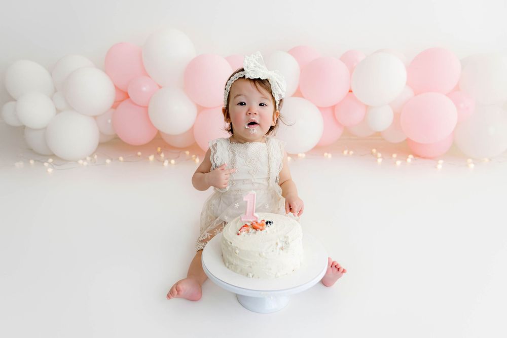 Baby girl in white dress sits with first birthday cake surrounded by pink and white balloons on white backdrop.