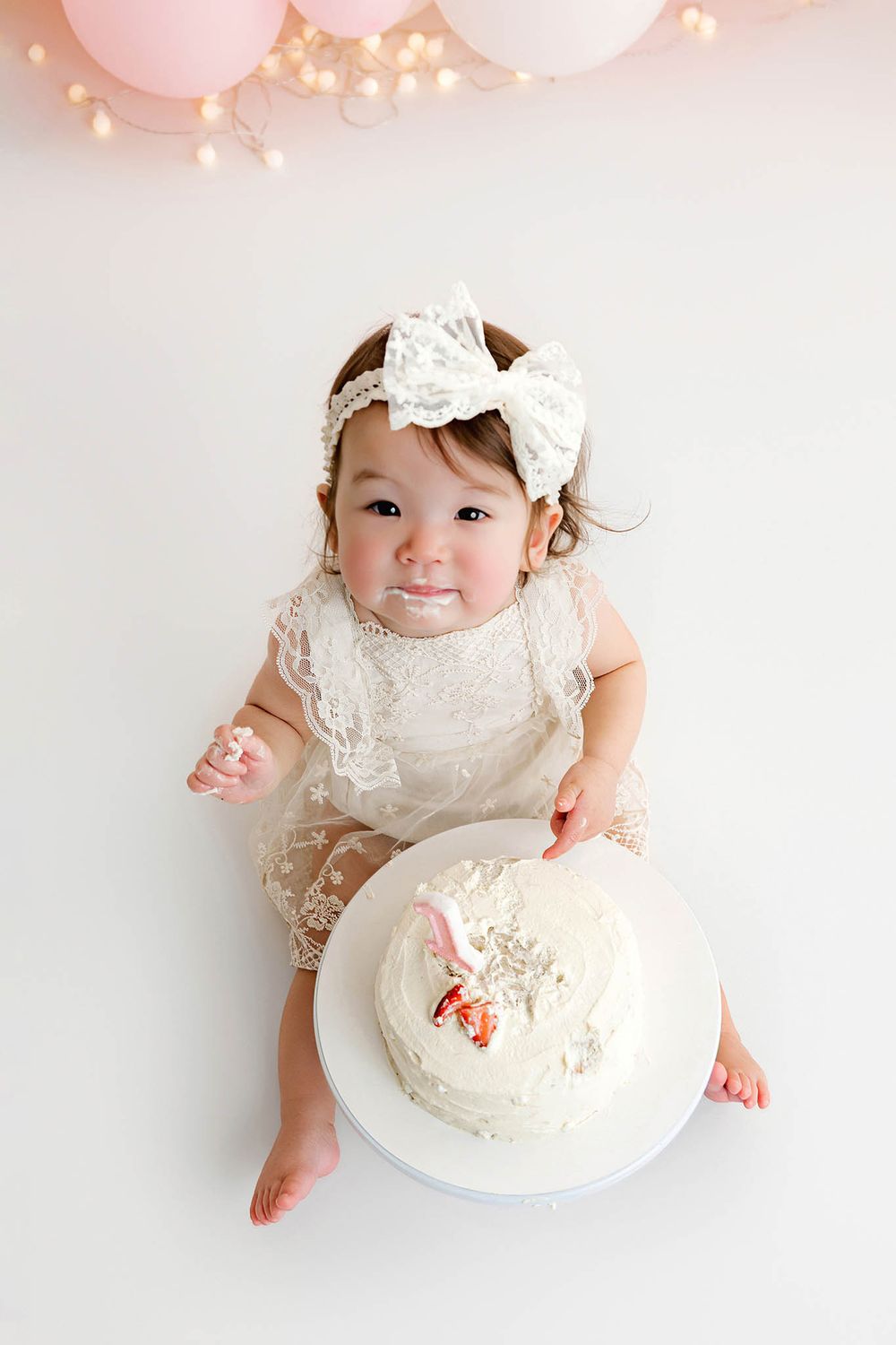 Baby girl in lace romper and bow headband smashing her first birthday cake, surrounded by pink balloons.